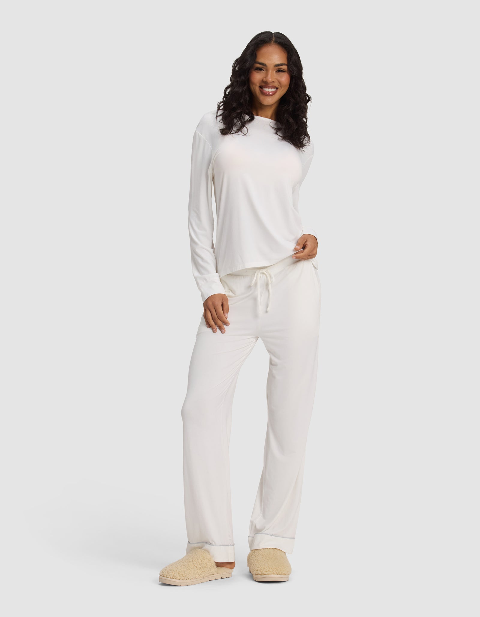A woman with long curly hair smiles while wearing the Cozy Earth Women's Bamboo Stretch-Knit Long Sleeve Lounge Tee, paired with beige slippers, against a plain light background.