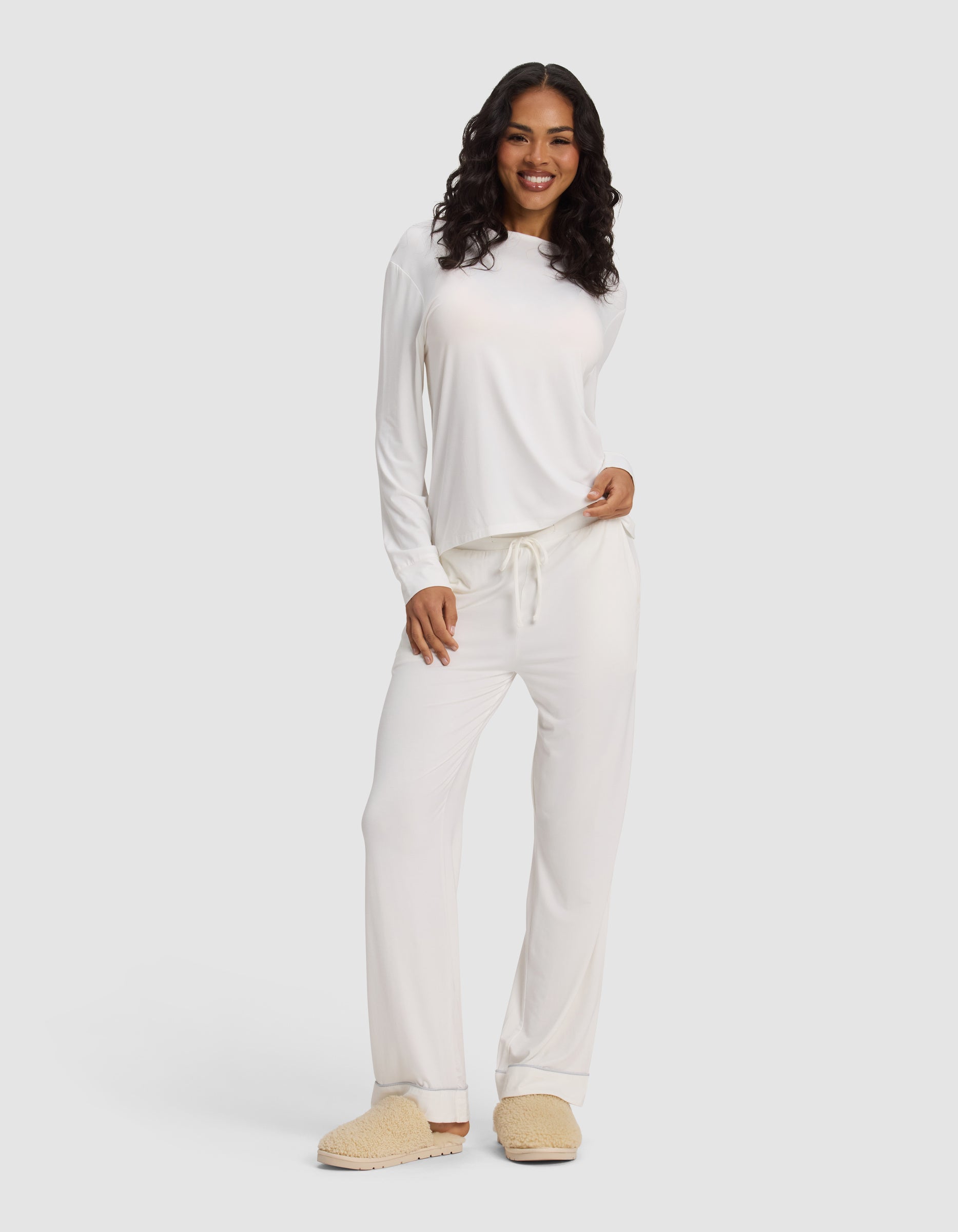 A woman with long curly hair smiles while wearing the Cozy Earth Women's Bamboo Stretch-Knit Long Sleeve Lounge Tee, paired with beige slippers, against a plain light background.