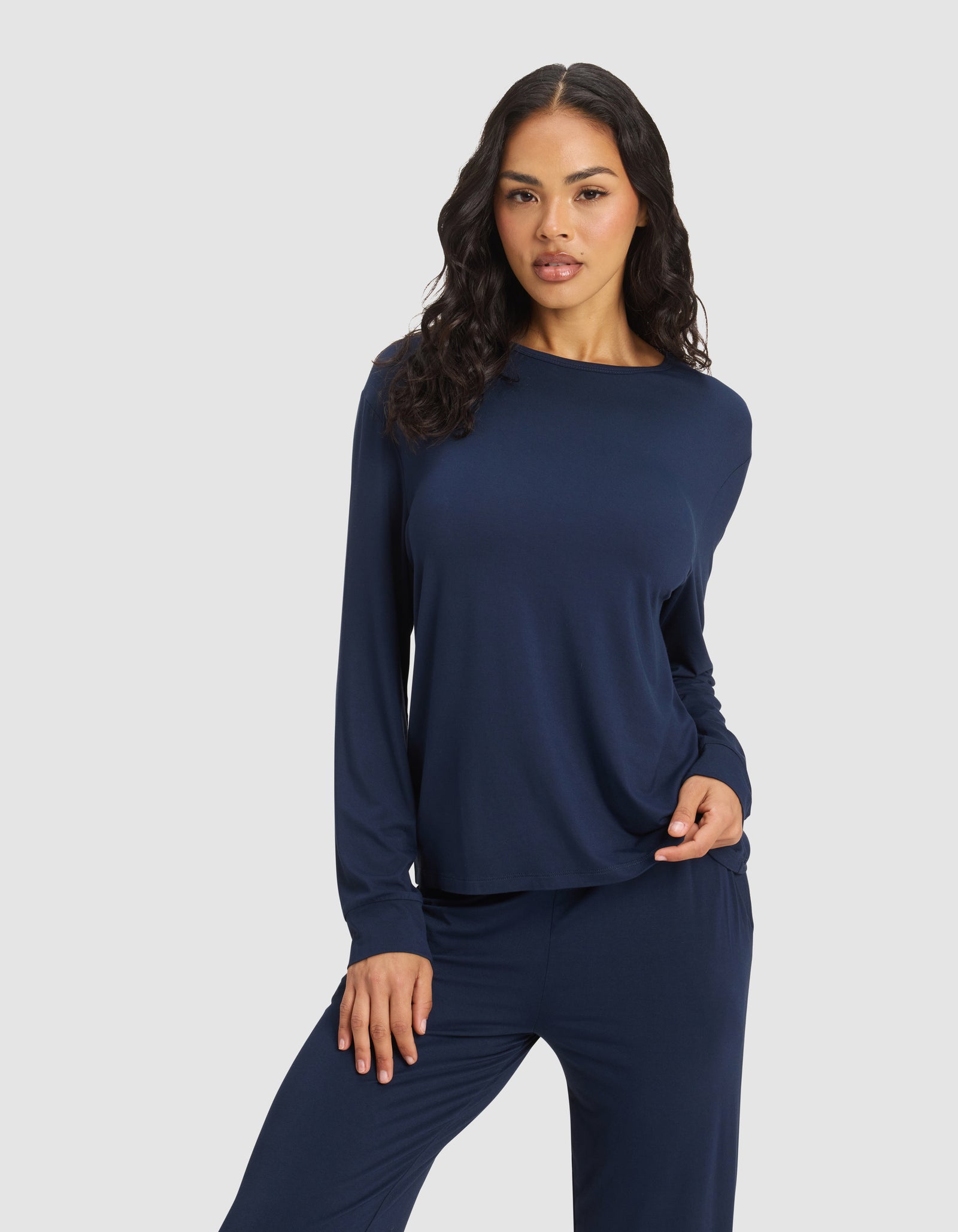 A woman with long, wavy dark hair models the Cozy Earth Women's Bamboo Stretch-Knit Long Sleeve Lounge Tee in navy blue, paired with matching pants, standing before a light gray background with one hand on her hip.
