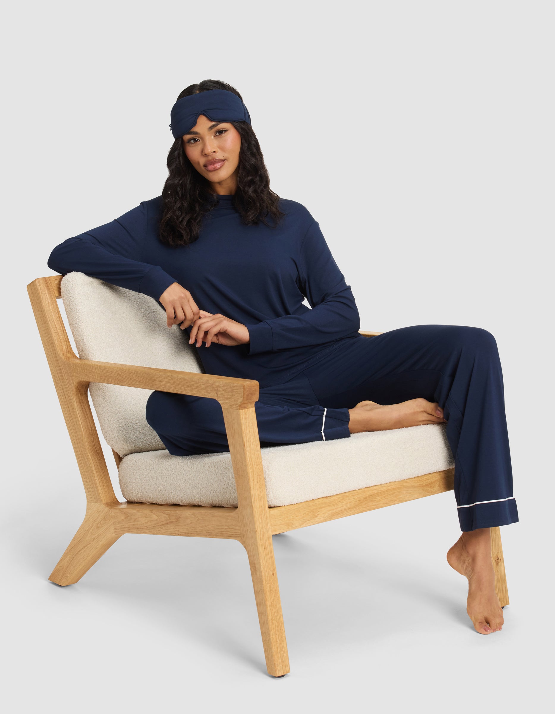 A woman relaxes barefoot on a cushioned wooden chair, wearing the Cozy Earth Women's Bamboo Stretch-Knit Long Sleeve Lounge Tee in navy blue with a matching sleep mask. One leg is folded up, arm resting on the back, set against a plain light background.