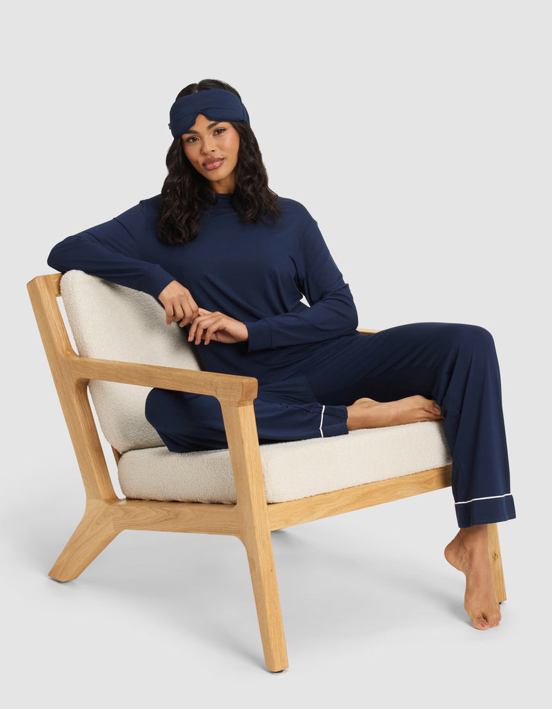 A woman relaxes barefoot on a cushioned wooden chair, wearing the Cozy Earth Women's Bamboo Stretch-Knit Long Sleeve Lounge Tee in navy blue with a matching sleep mask. One leg is folded up, arm resting on the back, set against a plain light background.