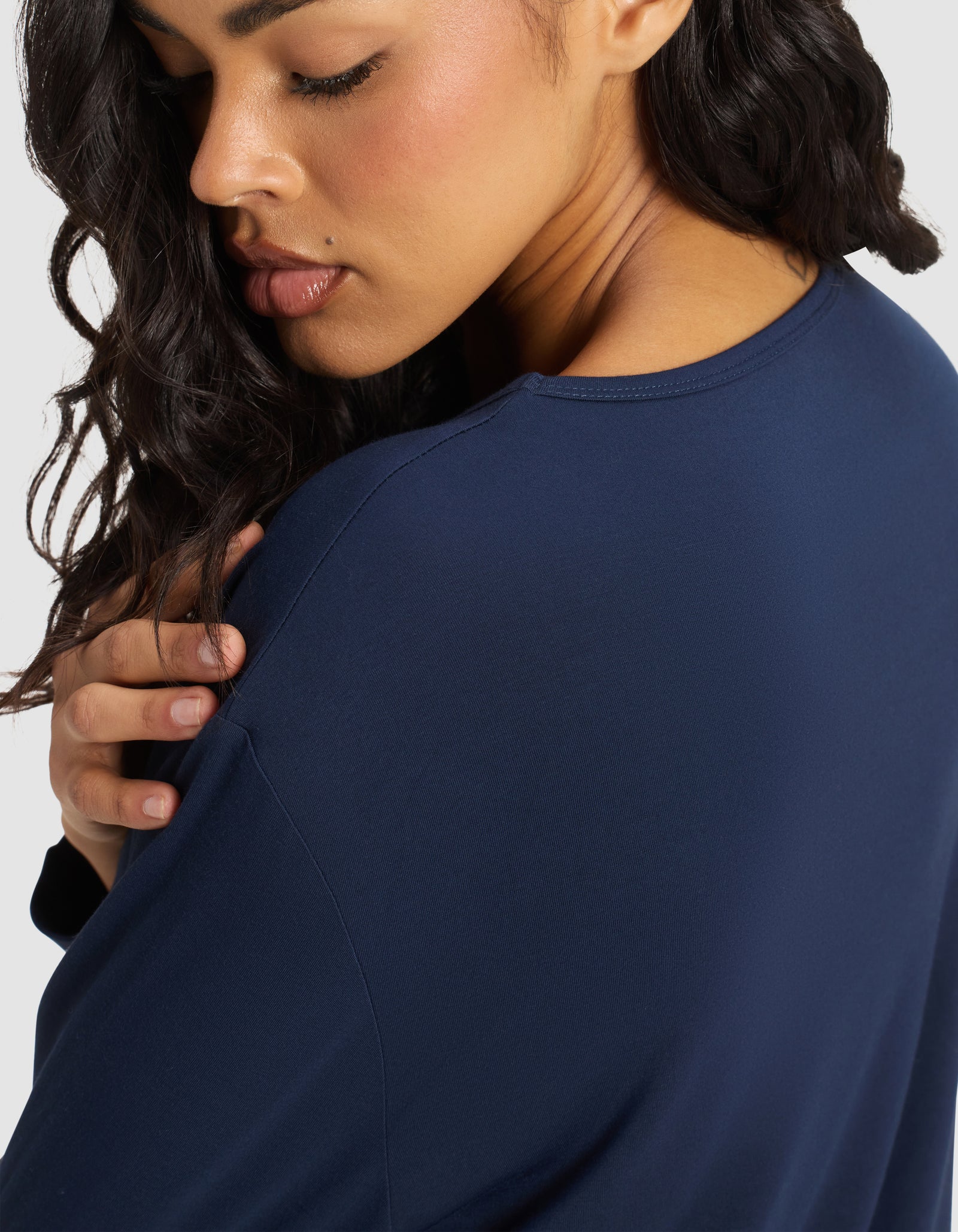A woman with wavy dark hair and glowing skin is shown in profile, gently touching her shoulder while wearing the Cozy Earth Women's Bamboo Stretch-Knit Long Sleeve Lounge Tee in navy against a plain background, her expression calm and thoughtful.