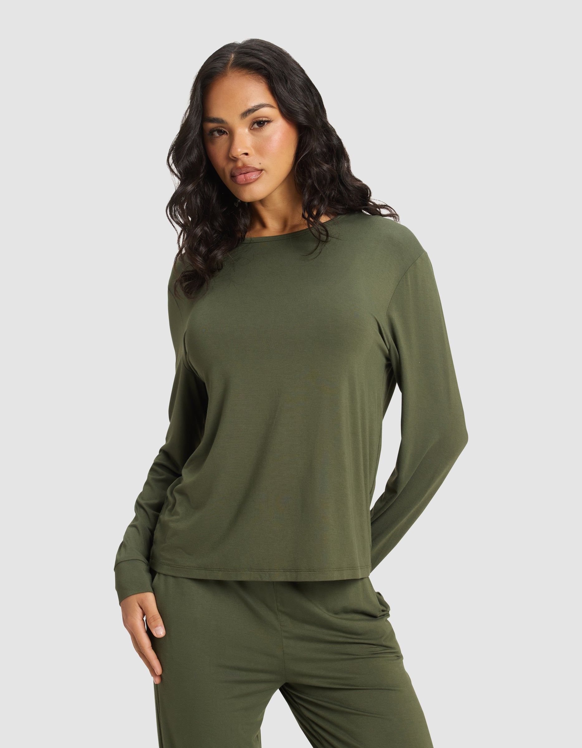 A woman with long, wavy dark hair wears the Cozy Earth Women's Bamboo Stretch-Knit Long Sleeve Lounge Tee in olive green, paired with matching pants, standing against a plain light gray background.