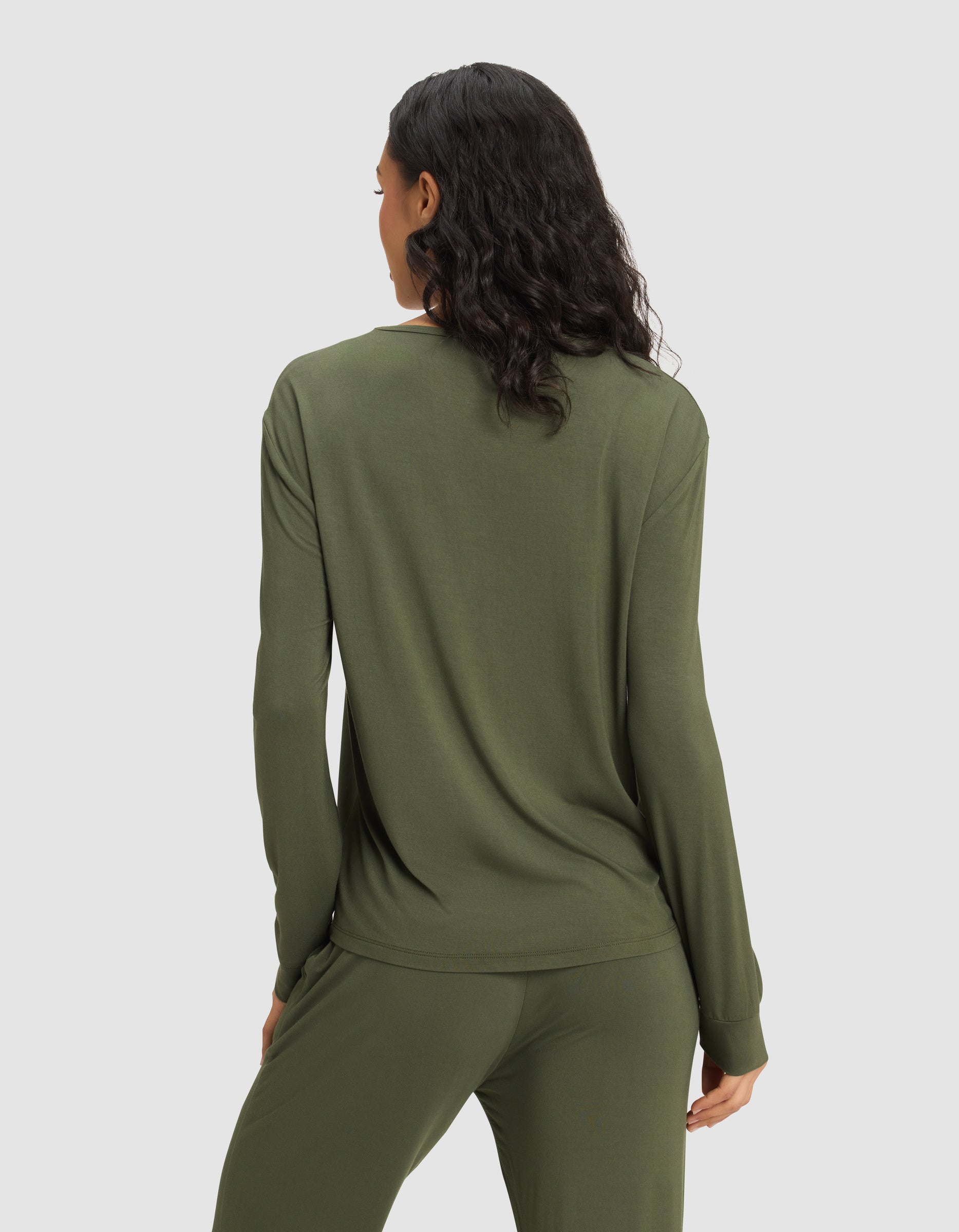 A woman with wavy dark hair is seen from behind, modeling Cozy Earth's Women's Bamboo Stretch-Knit Long Sleeve Lounge Tee in olive green against a plain light gray background.|Color:Olive