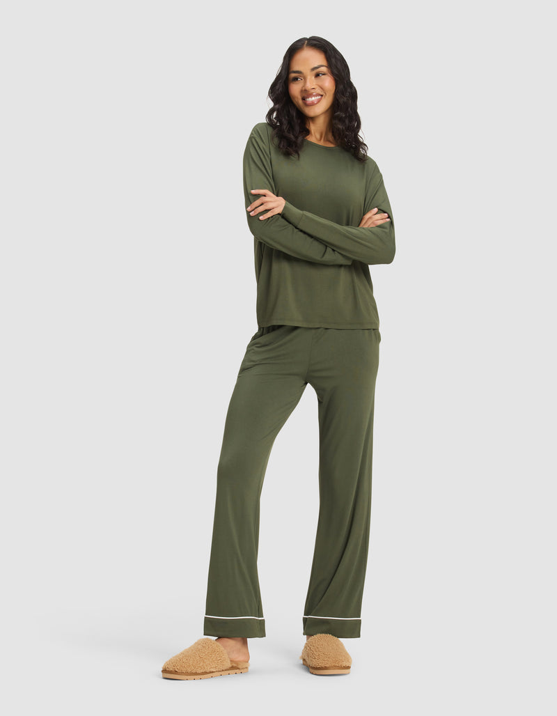 A woman smiles with arms crossed in Cozy Earth's Women's Bamboo Stretch-Knit Long Sleeve Lounge Tee, paired with matching olive green pants featuring white trim and tan fuzzy slippers, against a plain light gray background.