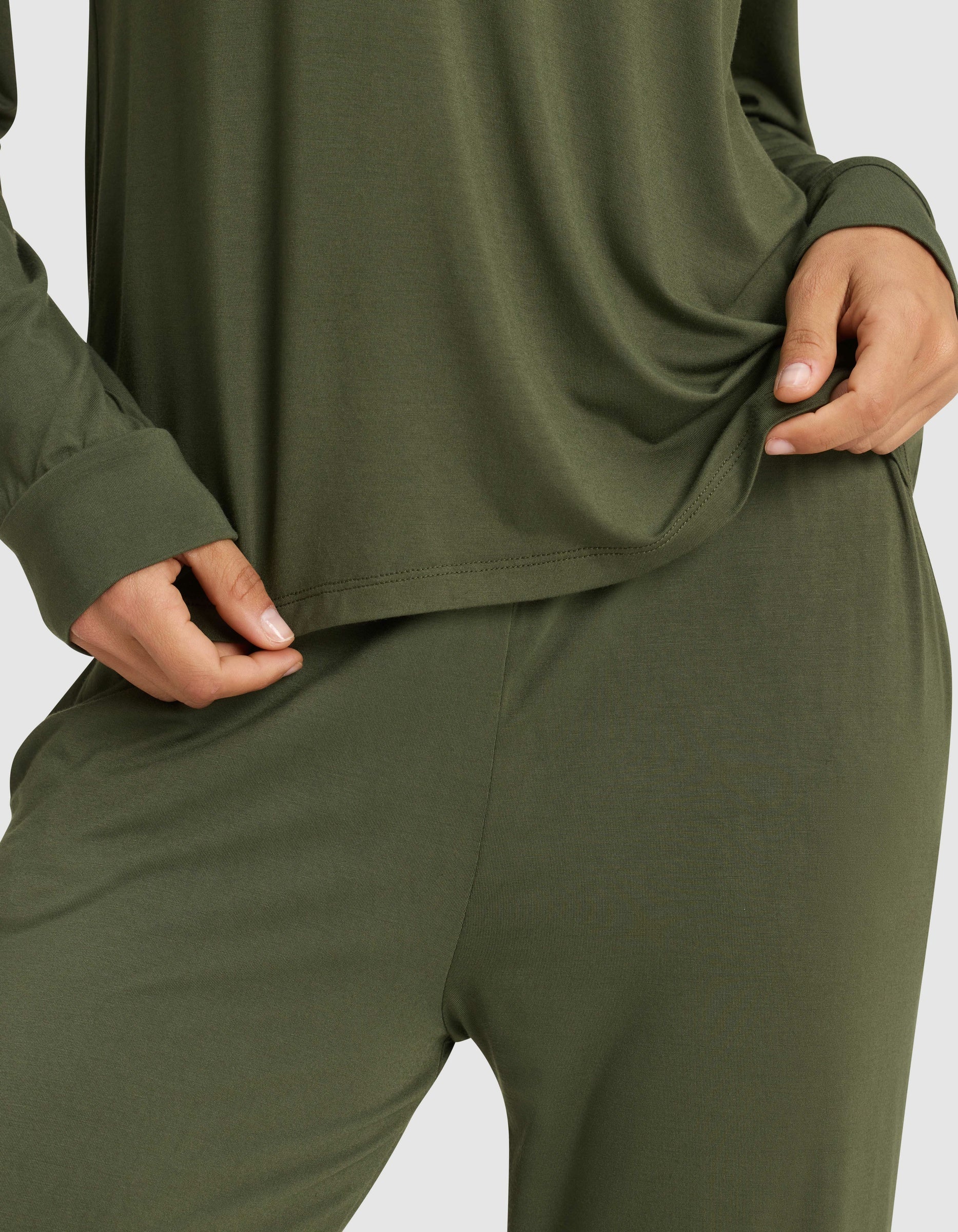 A woman wears the Cozy Earth Women's Bamboo Stretch-Knit Long Sleeve Lounge Tee in olive green, holding the shirt's hem; only her torso and upper legs are visible.|Color:Olive