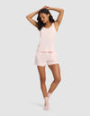 A woman models the Cozy Earth Women's Bamboo Stretch-Knit Tank & Short Set in light pink, paired with matching socks, posing with one arm raised against a plain light gray background.|Color:Blossom