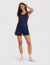 A woman poses against a white background in the Cozy Earth Women's Bamboo Stretch-Knit Tank & Short Set, navy blue, paired with light pink socks. She smiles with one hand to her head, showcasing long, wavy hair.|Color:Navy