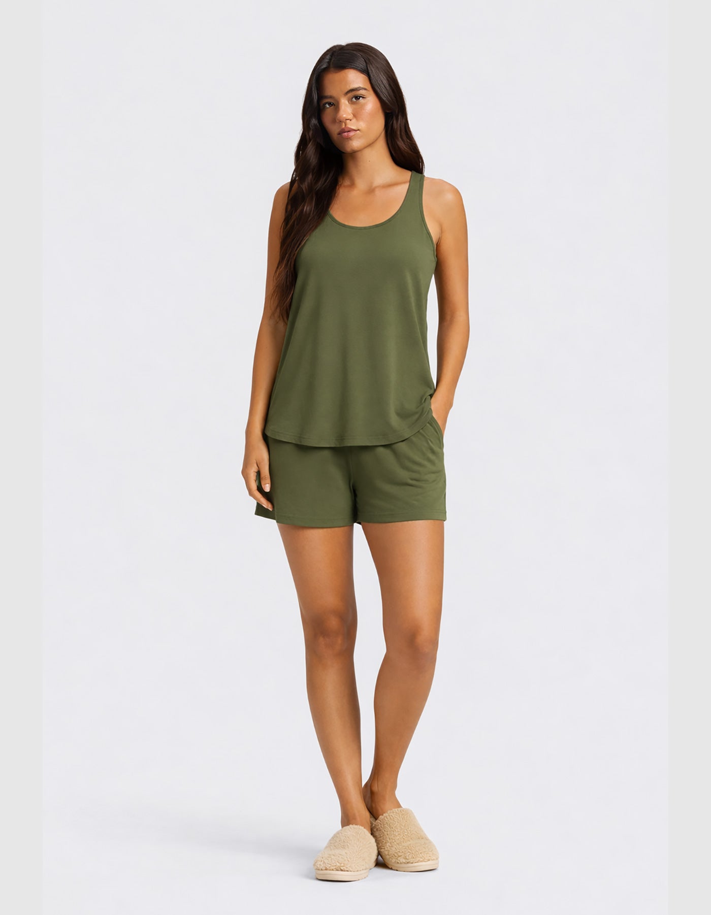 A woman faces forward in the Cozy Earth Women's Bamboo Stretch-Knit Tank & Short Set in olive green, paired with beige slippers. She has long straight hair, a neutral expression, and stands against a plain light background.