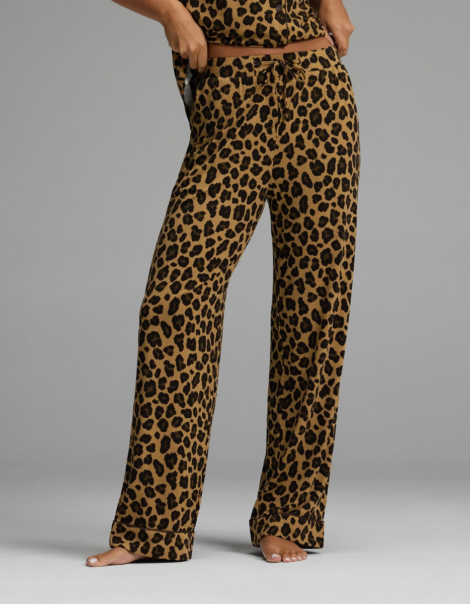 A person stands barefoot wearing Cozy Earth’s Women’s Bamboo Stretch-Knit Pajama Pant in a loose-fitting silhouette, featuring a brown base with a black leopard print. The image is cropped at the torso, concealing the individual’s upper body and face.