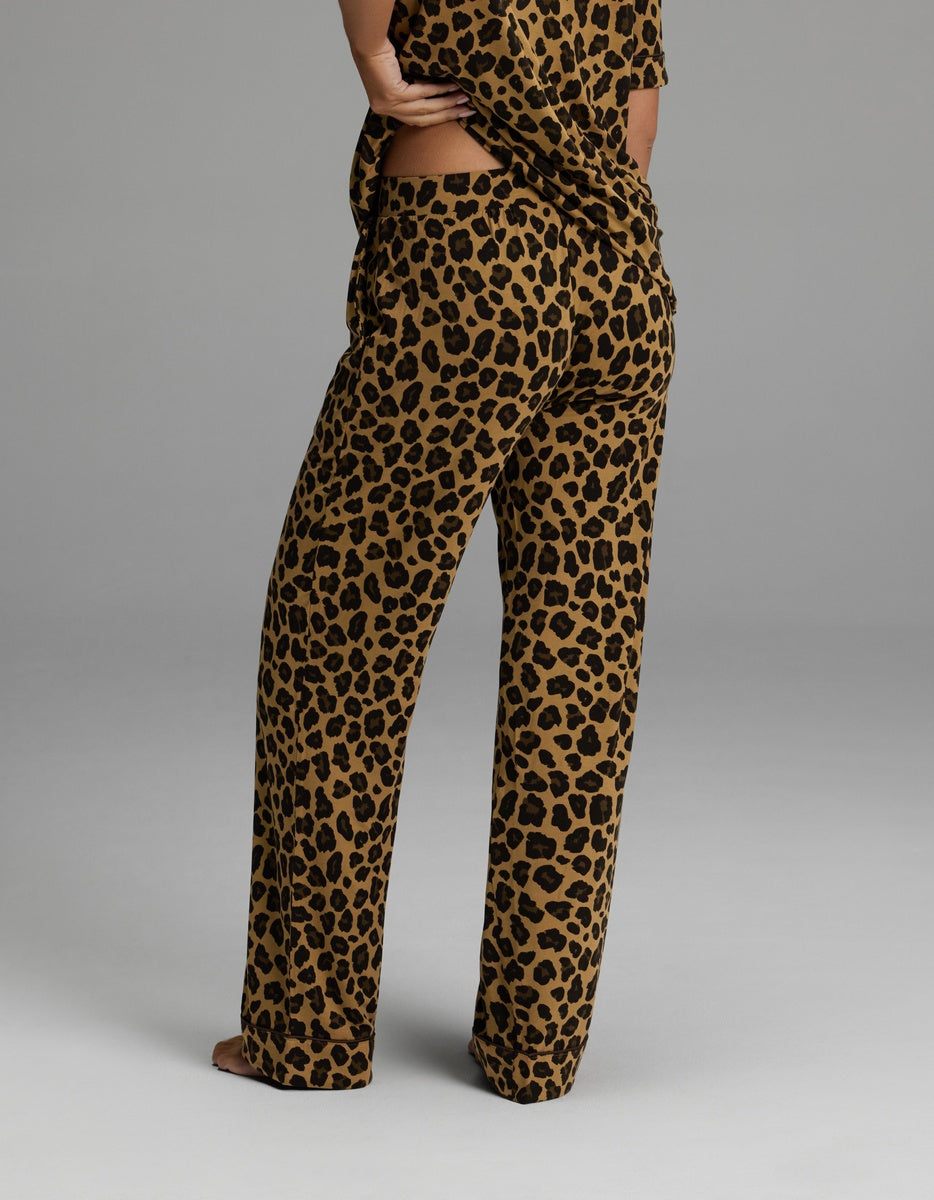 A person stands barefoot, shown from the waist down, wearing Cozy Earth’s Women’s Bamboo Stretch-Knit Pajama Pant in a loose-fitting leopard print with a matching top, against a plain grey background.