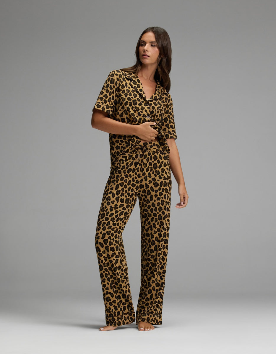 A woman stands barefoot against a plain gray background, wearing Cozy Earth’s Women’s Bamboo Stretch-Knit Pajama Pant in a matching leopard print set, featuring short sleeves and long pants.