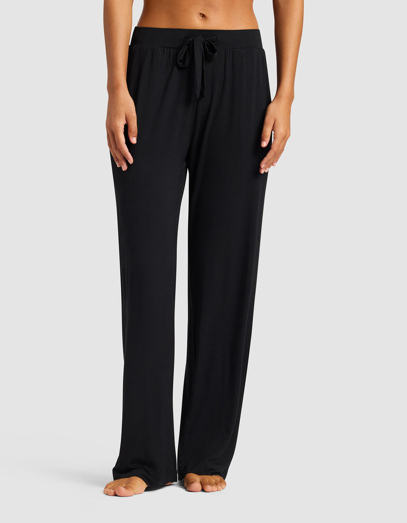 A woman stands barefoot against a plain light gray background, wearing Cozy Earth’s Women's Bamboo Stretch-Knit Pant in black with a drawstring waist and relaxed, comfortable fit.