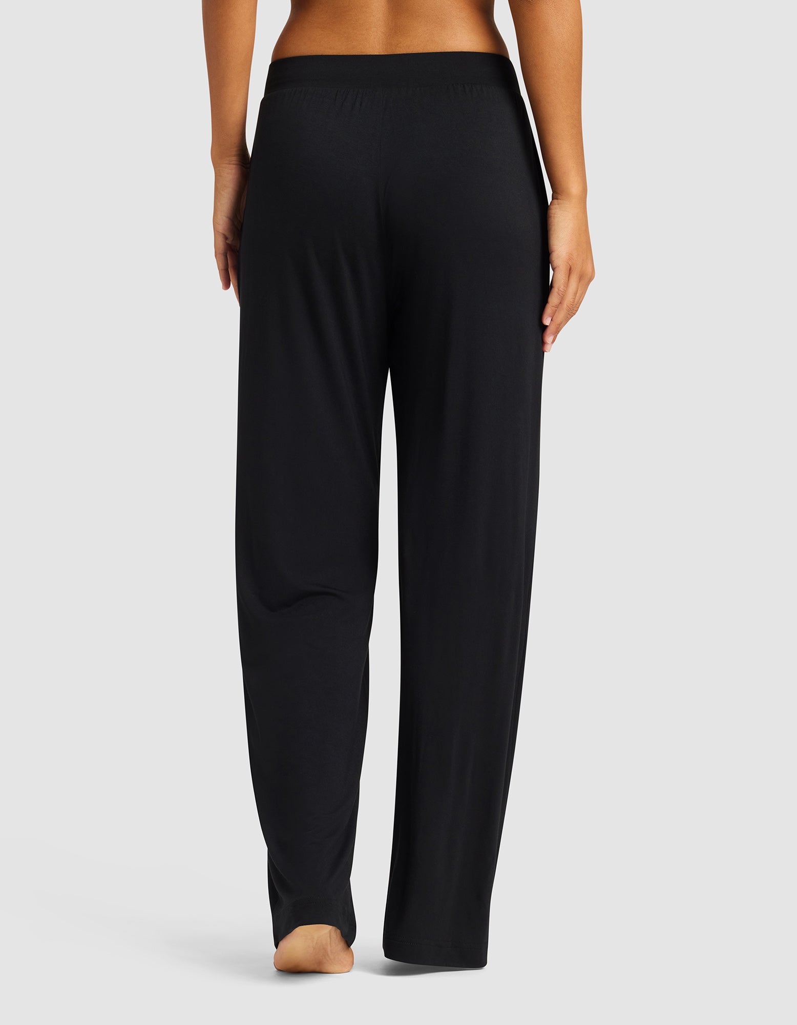 A person stands barefoot, facing away, wearing Cozy Earth’s Women's Bamboo Stretch-Knit Pant in black with an elastic waistband, set against a plain light gray background.