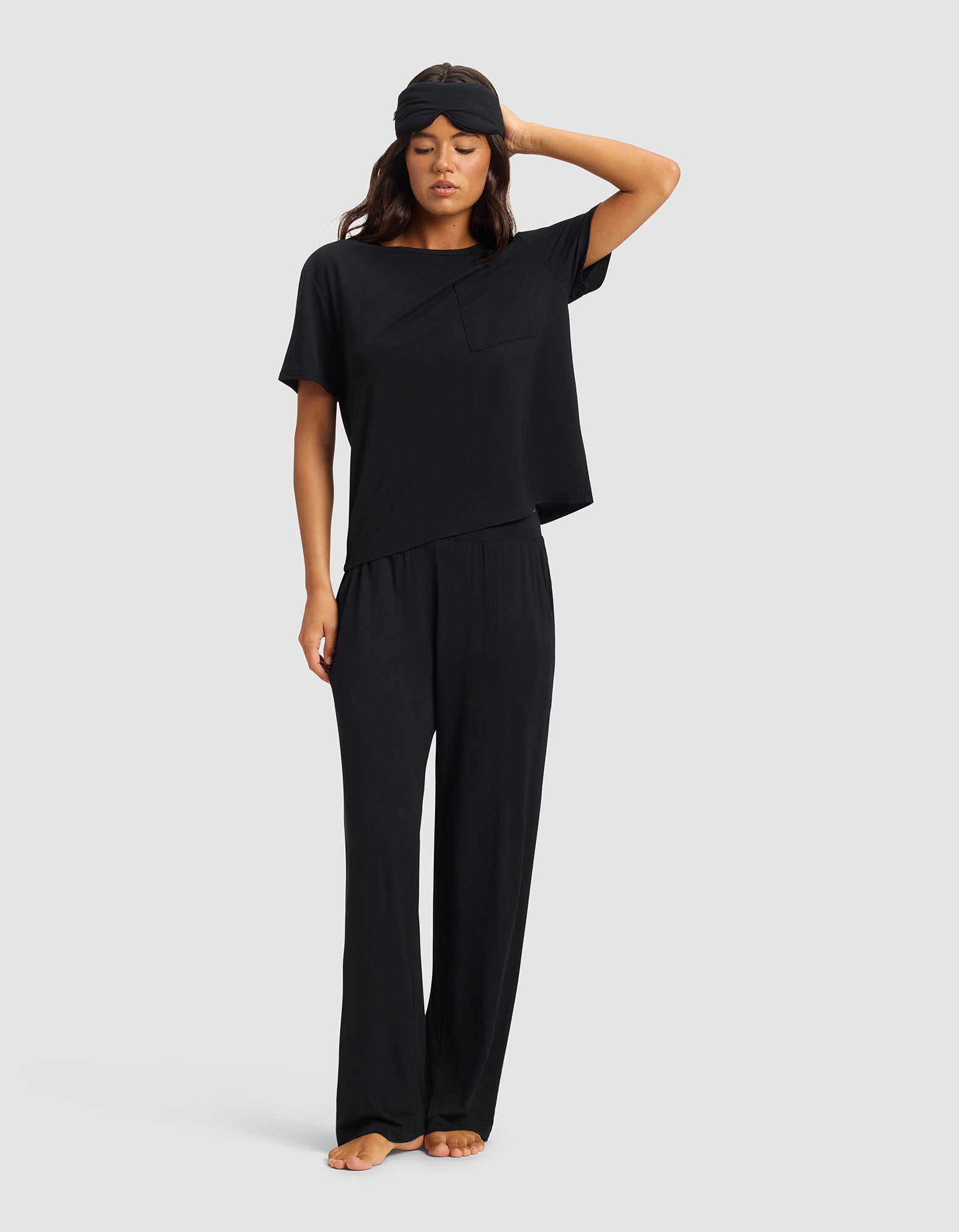 A woman stands barefoot, eyes closed, in Cozy Earth Women's Bamboo Stretch-Knit Pants with matching black pajamas and sleep mask, one hand touching her head, against a plain light background.
