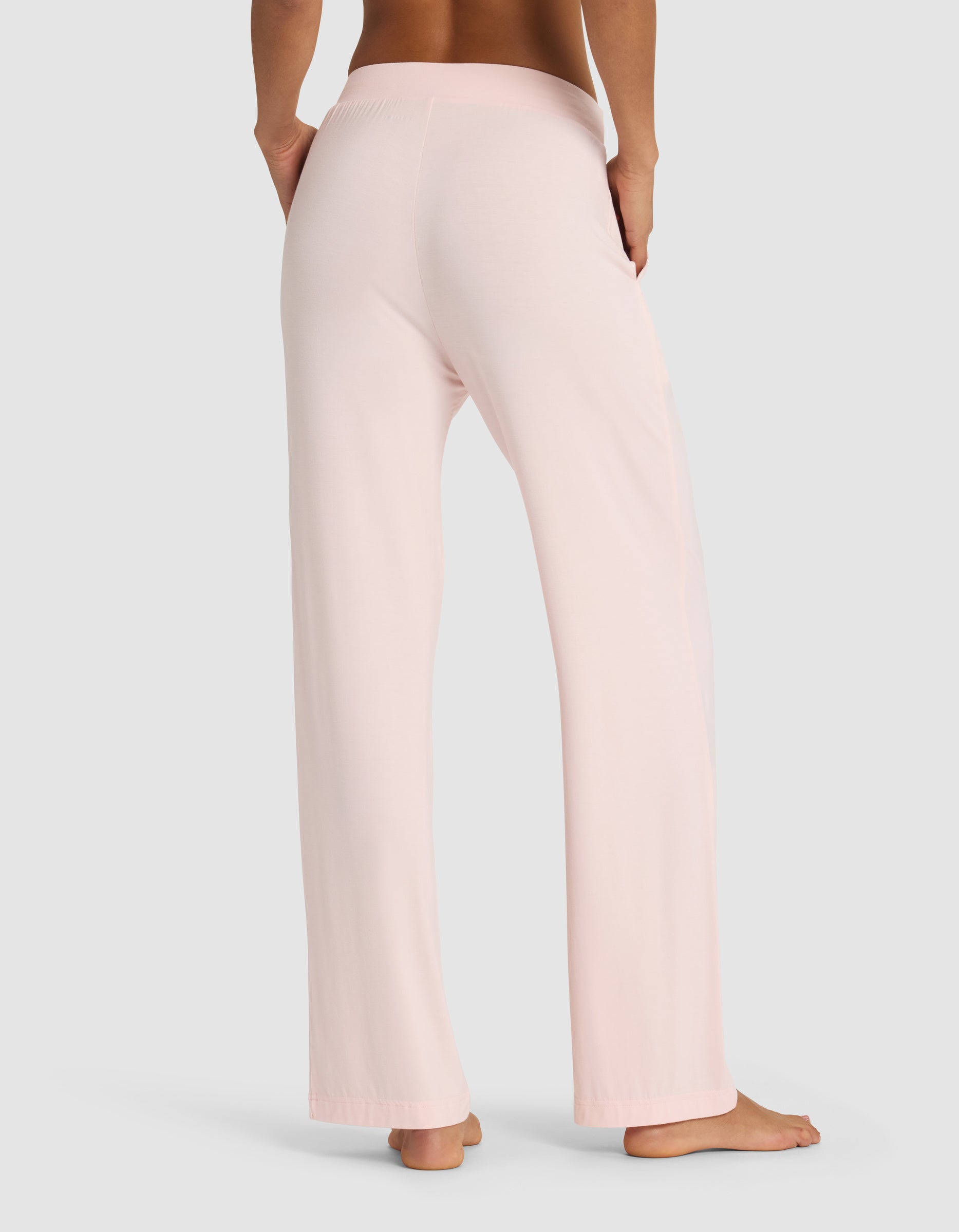 A person stands barefoot on a plain background, shown from the back with one hand on their hip, wearing Cozy Earth's Women's Bamboo Stretch-Knit Pant in light pink with a loose fit.|Color:Blossom