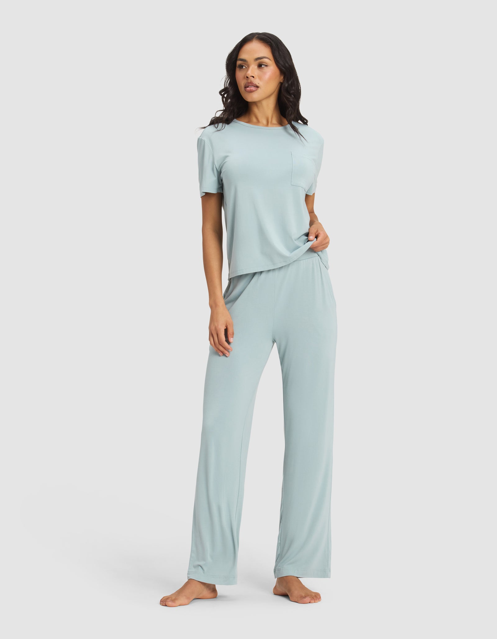A woman stands barefoot against a plain background, wearing Cozy Earth's Women's Bamboo Stretch-Knit Pant—a light blue, wide-leg pajama pant paired with a loose-fitting short-sleeve top.