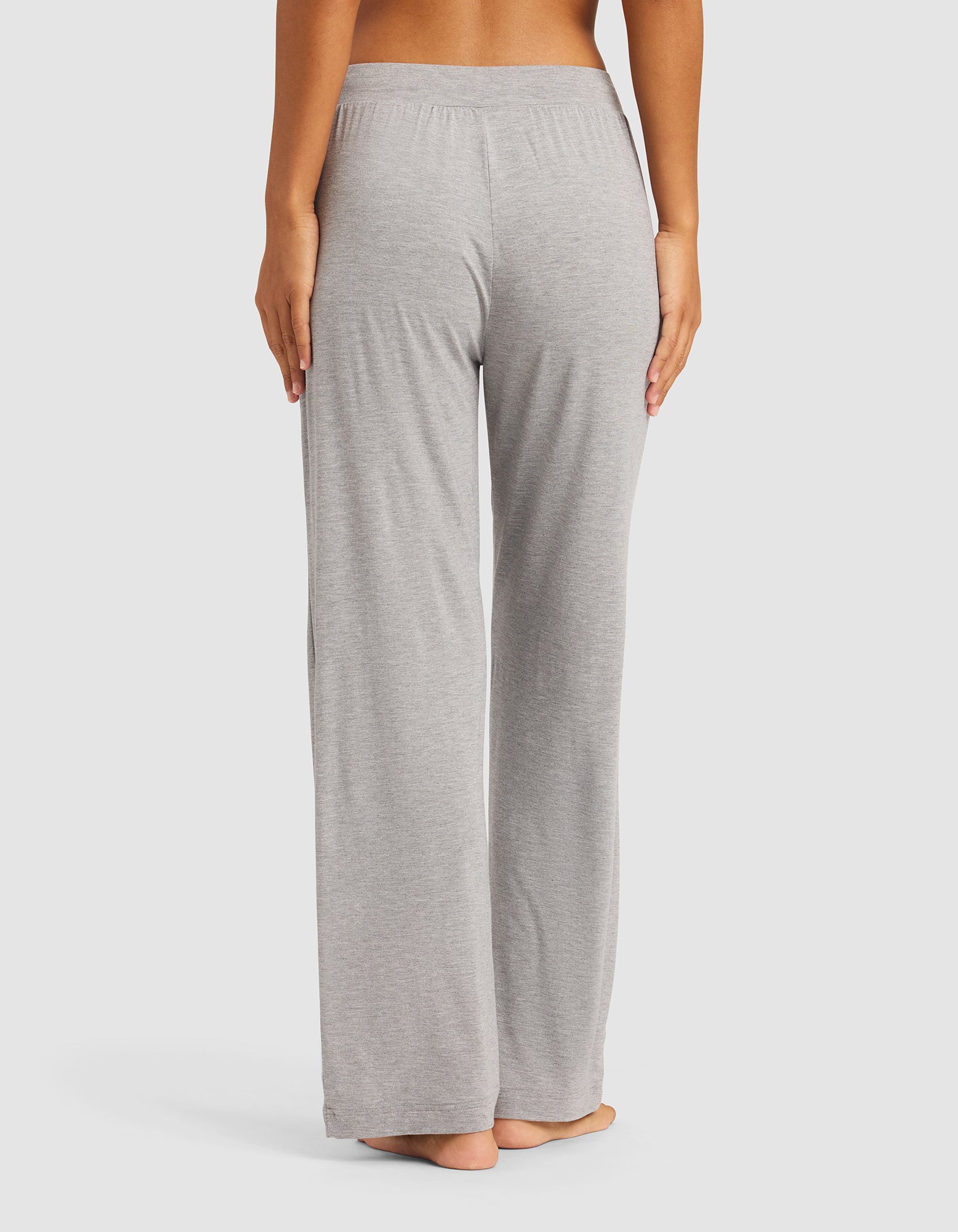 A person stands barefoot with their back to the camera, modeling Cozy Earth's Women's Bamboo Stretch-Knit Pant in light gray. The loose-fitting pants feature an elastic waistband, shown from the lower back down against a plain white background.|Color:Heather Grey