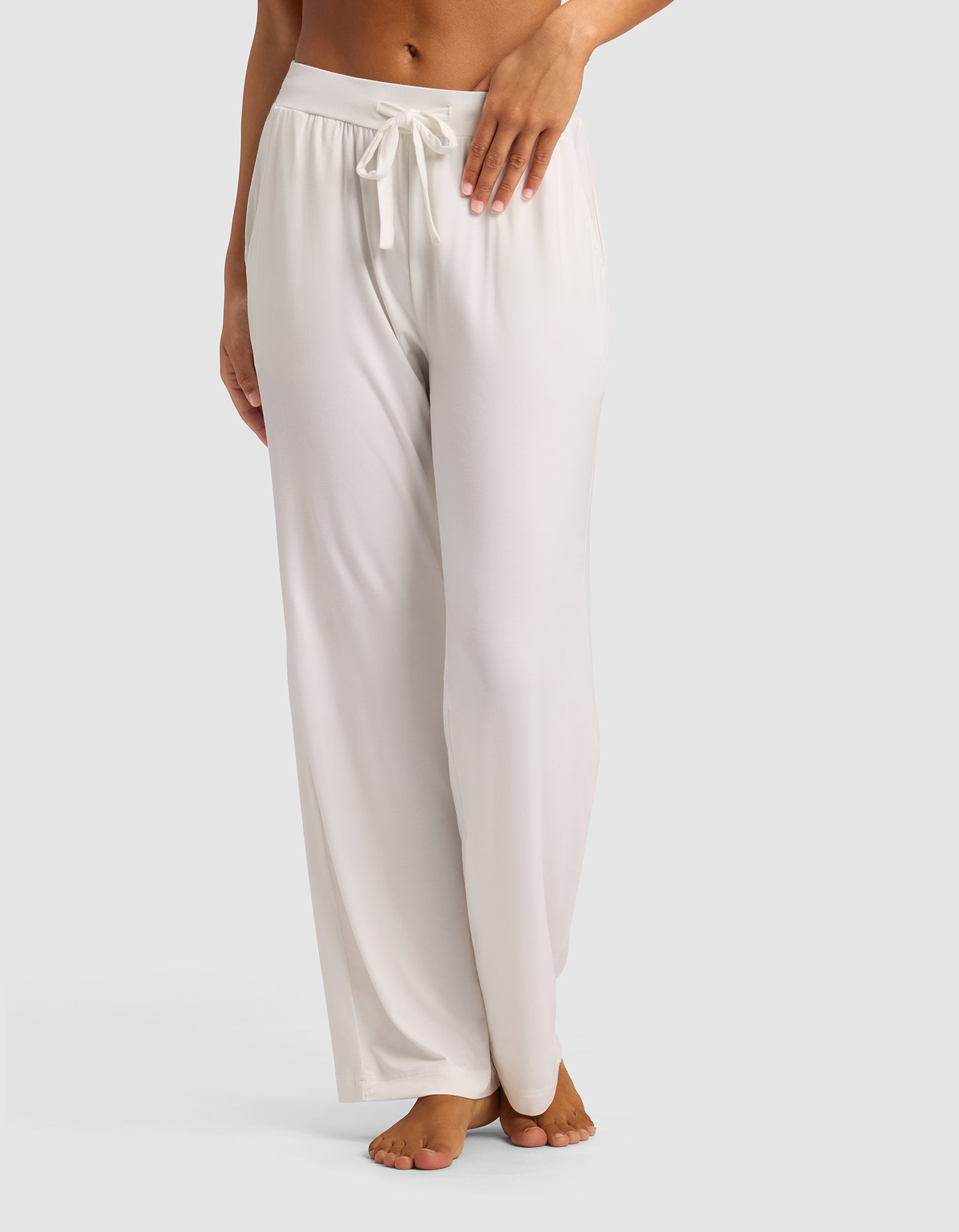 A woman stands barefoot against a plain light background, modeling the lower half of Cozy Earth's Women's Bamboo Stretch-Knit Pant—loose-fitting, light-colored lounge pants with a drawstring waist.