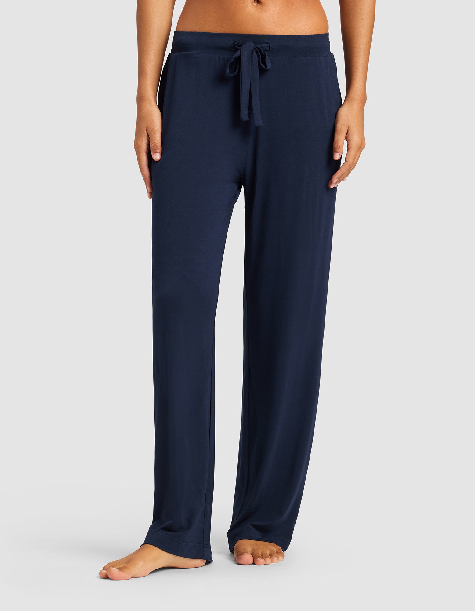 Model wears Cozy Earth Women’s Bamboo Stretch-Knit Pant in navy blue, featuring a loose fit and drawstring waist, shown barefoot with only the lower torso and legs visible against a light gray background.