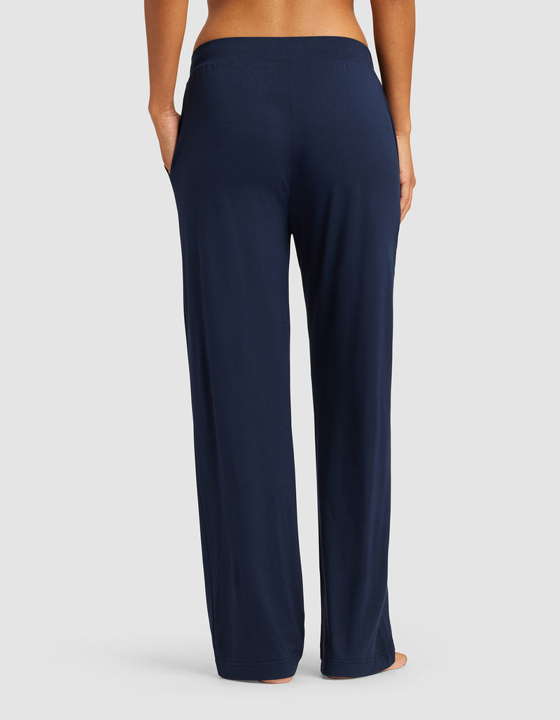 A woman is shown from the waist down, barefoot against a neutral background, wearing Cozy Earth Women's Bamboo Stretch-Knit Pant in navy blue with a relaxed fit and elastic waistband.