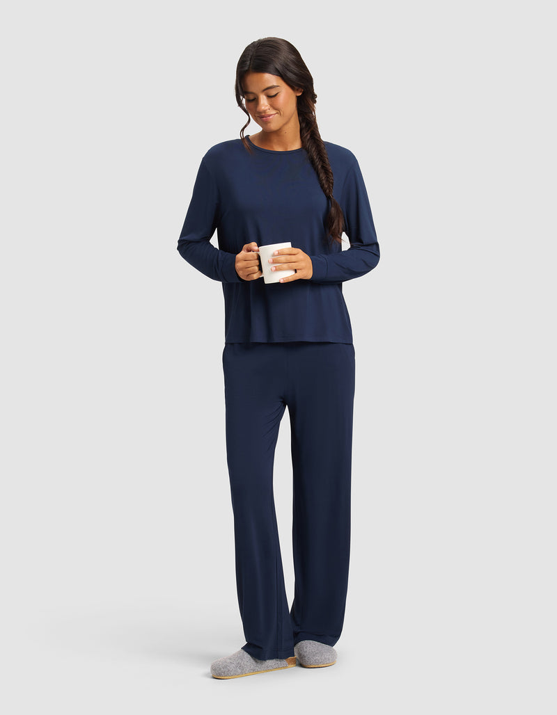 A woman with long dark hair in a braid wears Cozy Earth’s Women's Bamboo Stretch-Knit Pant in navy and gray slippers, holds a white mug, and smiles while looking down against a plain light gray background.