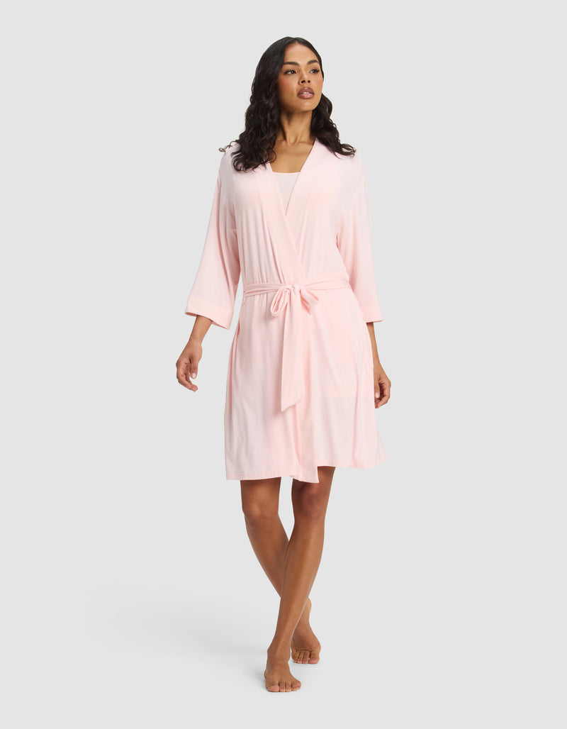 A woman wears the Cozy Earth Women's Bamboo Stretch-Knit Kimono Robe in light pink, knee-length with a belt, standing barefoot against a plain light gray background.