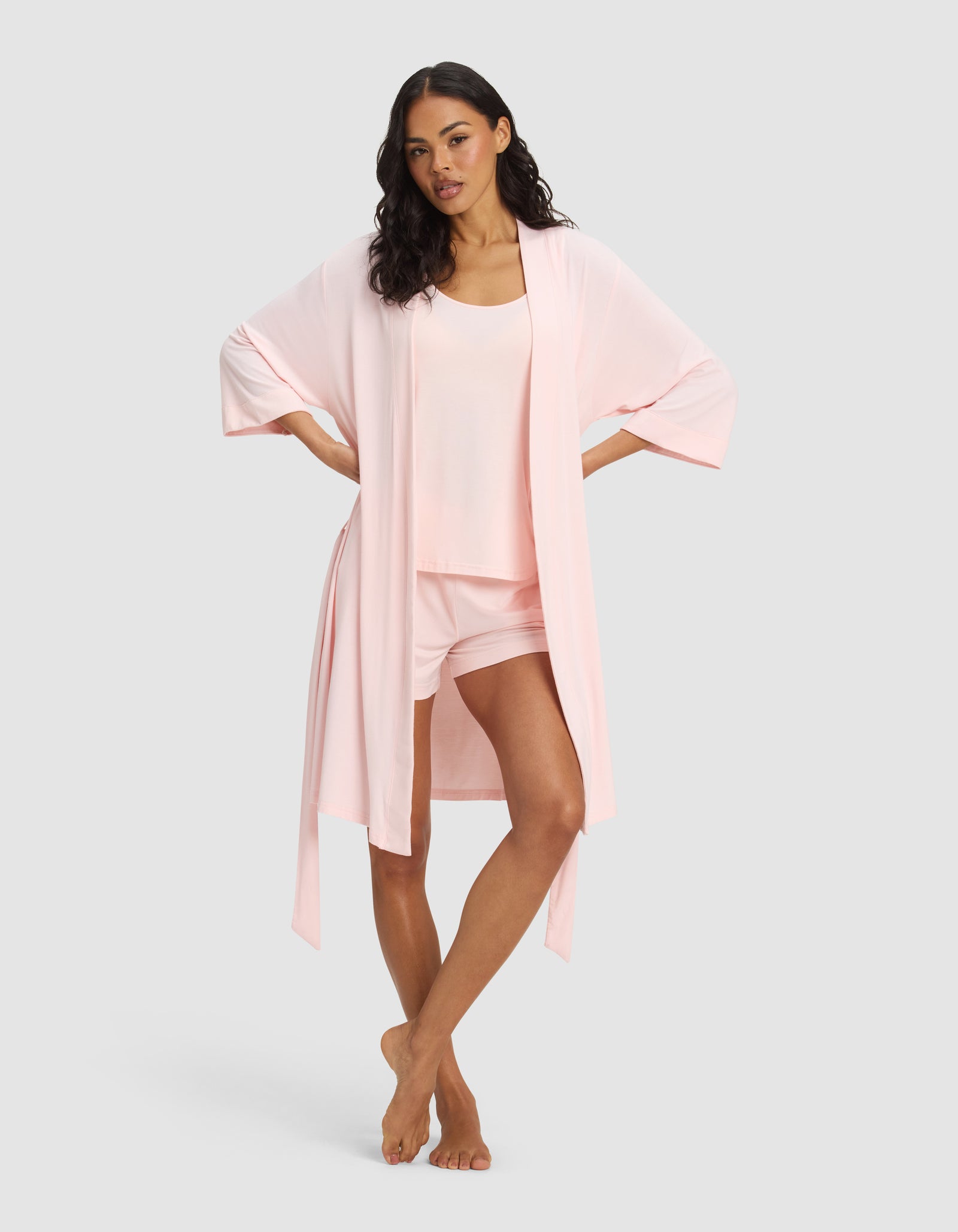 A woman stands barefoot against a gray background, wearing the Cozy Earth Women's Bamboo Stretch-Knit Kimono Robe—a light pink, open-front robe with wide sleeves—over a matching tank and shorts loungewear set.