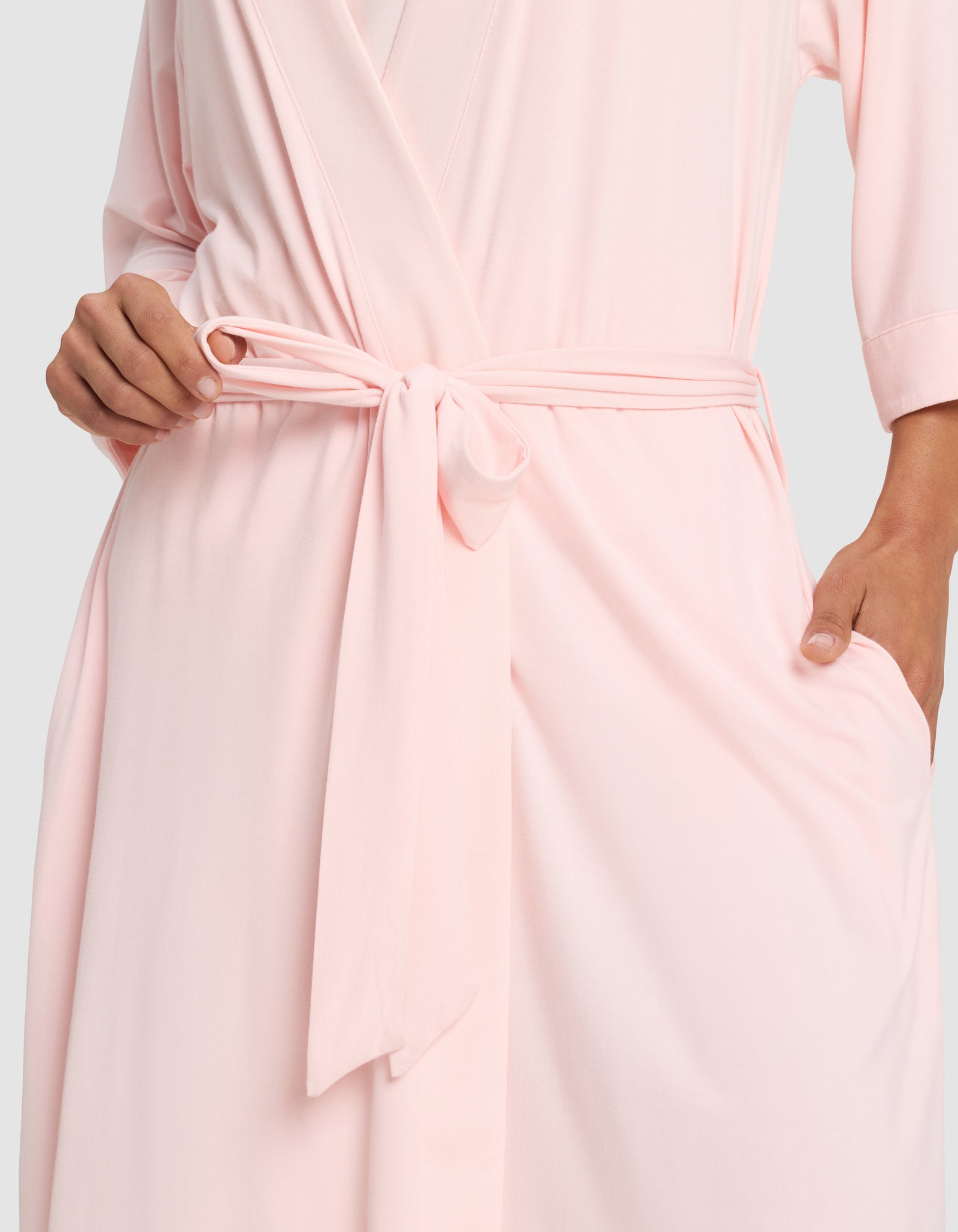 A woman models the Cozy Earth Women's Bamboo Stretch-Knit Kimono Robe in light pink, tied at the waist with a matching sash, with one hand on her hip and the other touching the sash.
