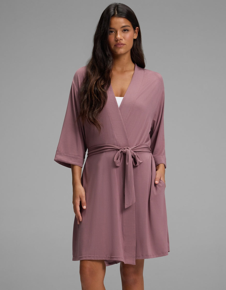 A woman wearing a mauve knee-length Women’s Bamboo Stretch-Knit Kimono Robe by Cozy Earth with a tie waist and elbow-length sleeves.