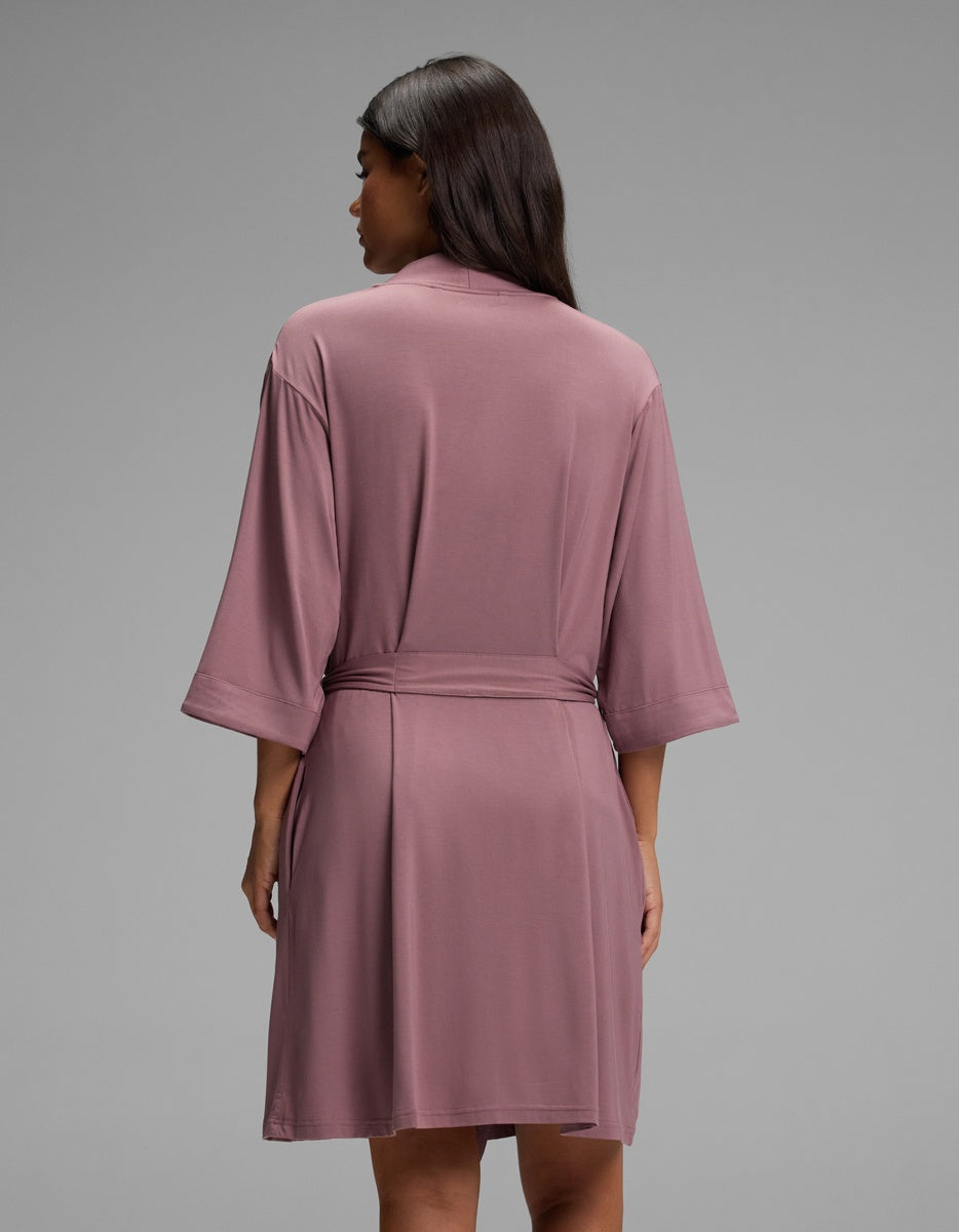 A woman seen from behind wearing a mauve Cozy Earth Women’s Bamboo Stretch-Knit Kimono Robe with a tie waist and elbow-length sleeves.