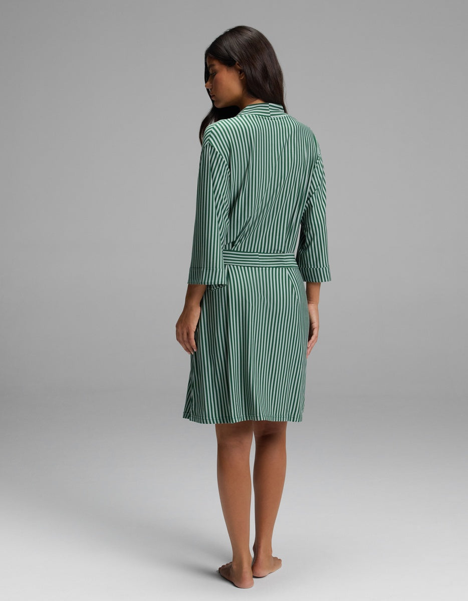 A woman wearing a knee-length, green and white striped Cozy Earth Women’s Bamboo Stretch-Knit Kimono Robe with three-quarter sleeves and a tied waist belt.