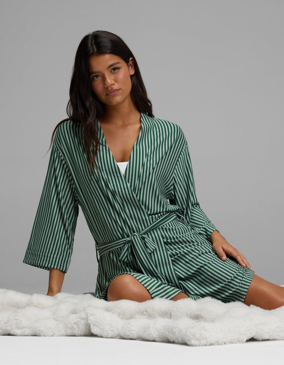 A woman wearing a knee-length green and white striped Women’s Bamboo Stretch-Knit Kimono Robe with a tie waist, sitting on a plush white surface.