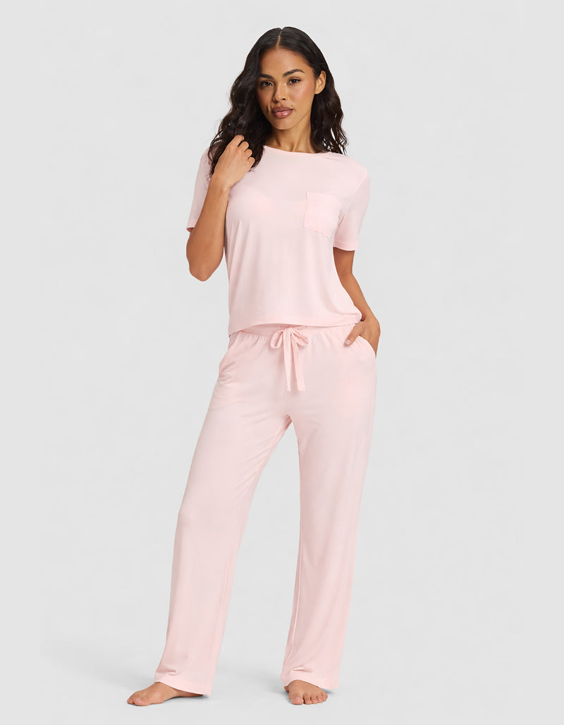 A woman poses barefoot against a plain gray background, wearing Cozy Earth’s Women's Bamboo Stretch-Knit Lounge Tee & Pant Set in light pink with short sleeves, pockets, and drawstring pants.