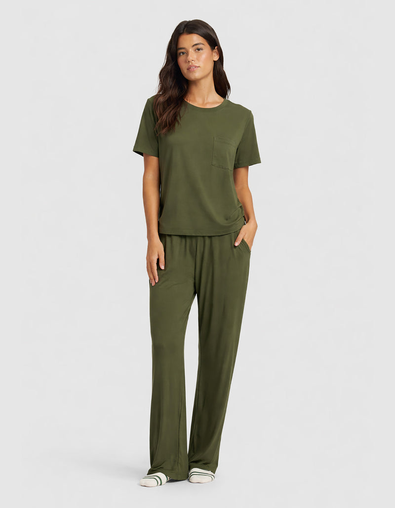 A woman wears the Cozy Earth Women's Bamboo Stretch-Knit Lounge Tee & Pant Set in olive green, featuring a chest pocket and loose pants, accessorized with white sneakers with green stripes.