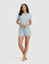 A woman stands barefoot against a light background, wearing the Cozy Earth Women's Bamboo Stretch-Knit Lounge Tee & Short Set in light blue. Her loose, wavy hair and gently resting arms complete the relaxed look.|Color:Brook
