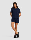 A woman poses against a plain background wearing the Cozy Earth Women's Bamboo Stretch-Knit Lounge Tee & Short Set in navy, paired with black slippers and her hands in her pockets.|Color:Navy