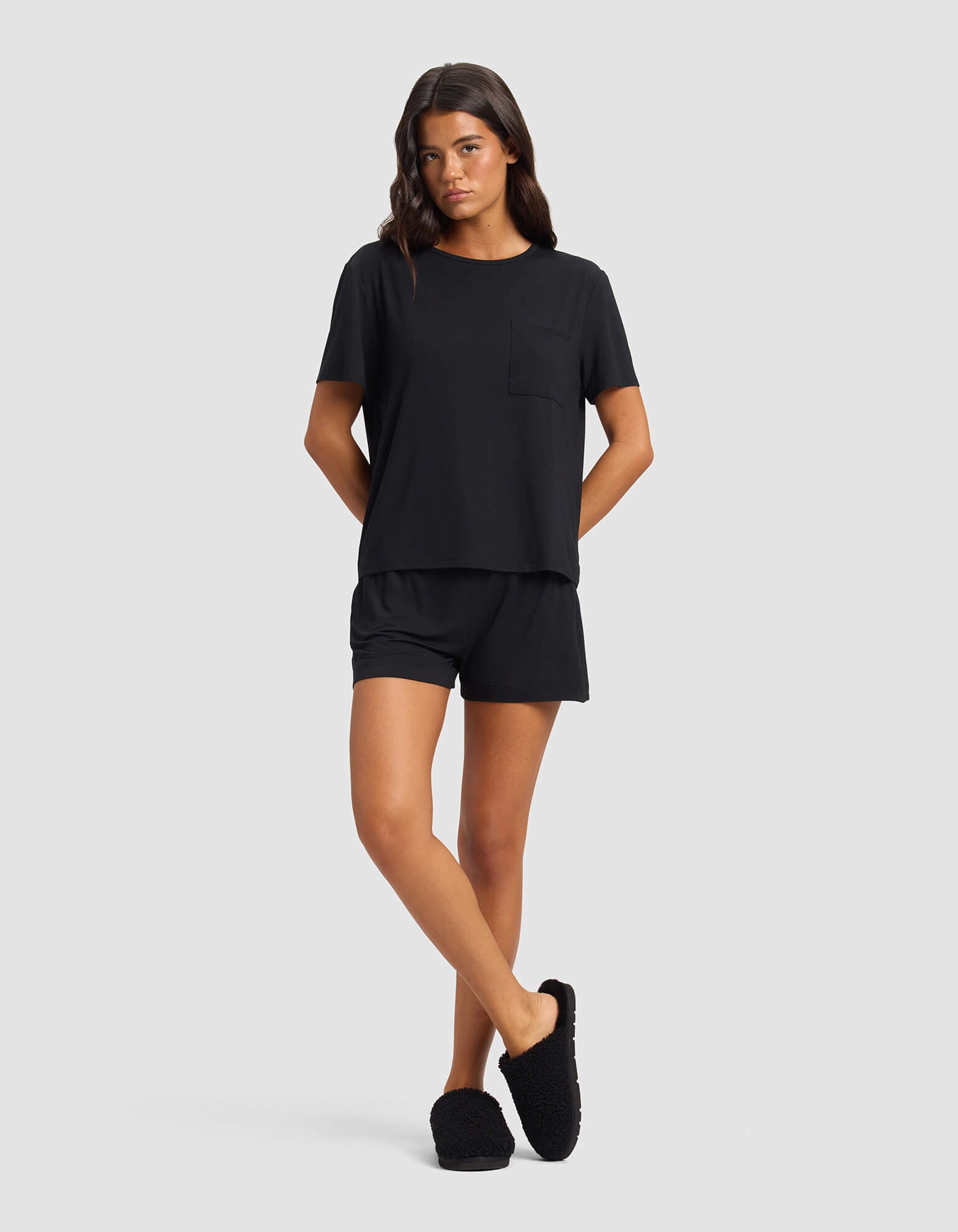 A woman wears the Cozy Earth Women's Bamboo Stretch-Knit Lounge Tee & Short Set in black, featuring a short-sleeve top with chest pocket and matching shorts. She stands against a plain background with hands behind her back and a neutral expression.|Color:Black