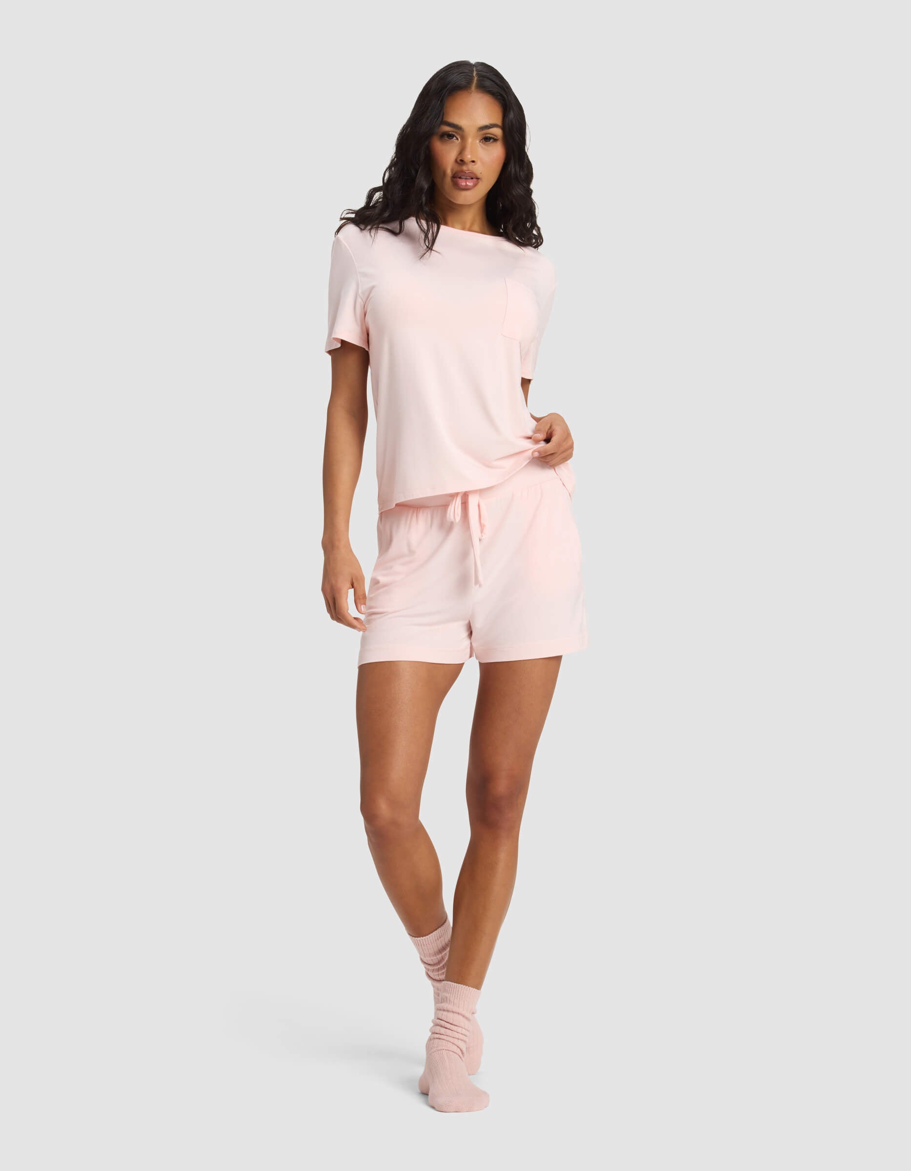 A woman wearing the Cozy Earth Women's Bamboo Stretch-Knit Lounge Tee & Short Set in light pink stands against a grey background, one hand on her waist, looking confidently at the camera.