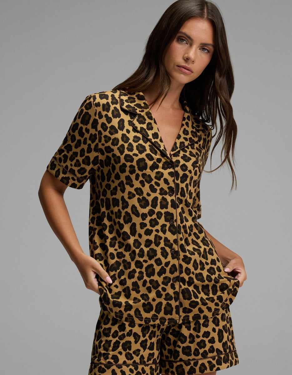 A woman stands against a plain background wearing the Cozy Earth Women’s Bamboo Stretch-Knit Short Sleeve Pajama Top paired with matching shorts, both featuring a bold brown and black leopard print pattern.