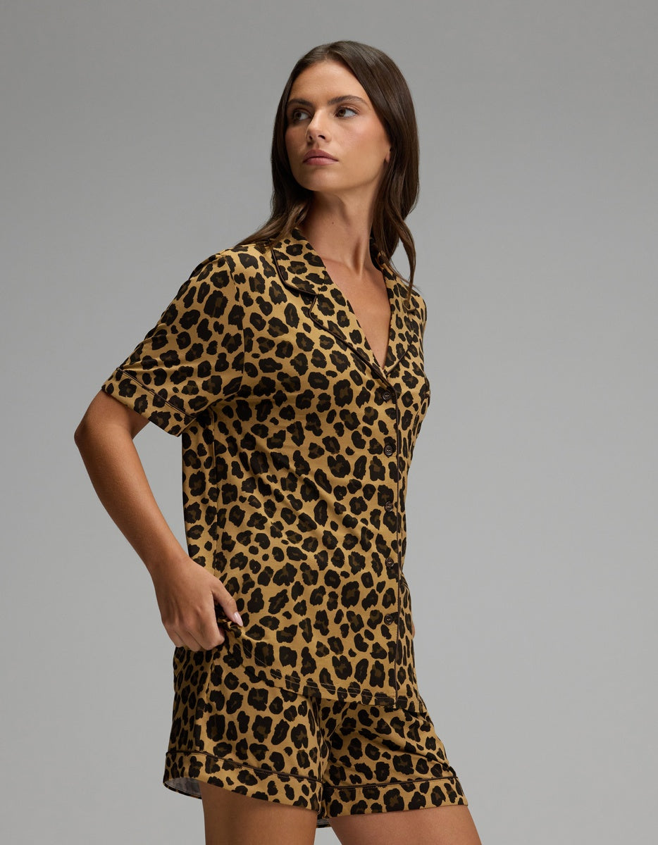 A woman wearing the Cozy Earth Women’s Bamboo Stretch-Knit Short Sleeve Pajama Set, featuring a short-sleeve, button-up top and matching pants in a brown and black leopard print pattern, stands against a plain gray background and looks to the side.