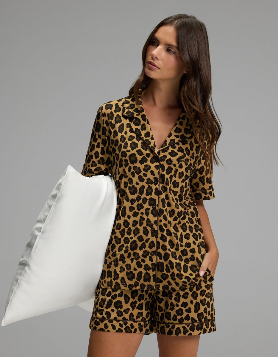 A woman wearing the Cozy Earth Women’s Bamboo Stretch-Knit Short Sleeve Pajama Set holds a large white pillow under her arm, standing against a plain gray background.