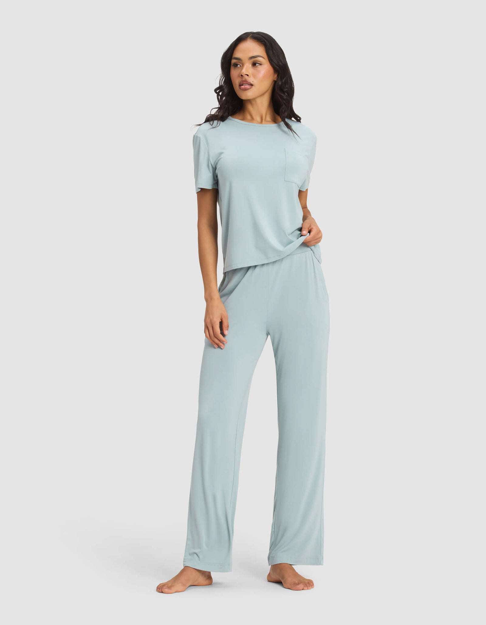 A woman stands barefoot against a plain light background, wearing the Cozy Earth Women's Bamboo Stretch-Knit Lounge Tee & Pant Set in light blue. She holds the hem of the top and looks slightly to the side.