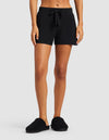 A person stands on a gray background wearing Cozy Earth Women's Bamboo Stretch-Knit Shorts in black. Their arms are relaxed by their sides, and their head is not visible in the image.|Color:Black