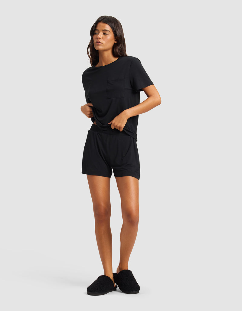 A woman with medium-length dark hair poses against a light gray background, wearing Cozy Earth's Women's Bamboo Stretch-Knit Shorts in black, a matching t-shirt, and slippers. She stands relaxed, holding the hem of her shirt.