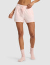 A person models Cozy Earth's Women's Bamboo Stretch-Knit Shorts in light pink, paired with matching socks, standing against a plain light grey background. The image highlights the shorts' fit from lower torso to legs, with one arm bent at the elbow.|Color:Blossom
