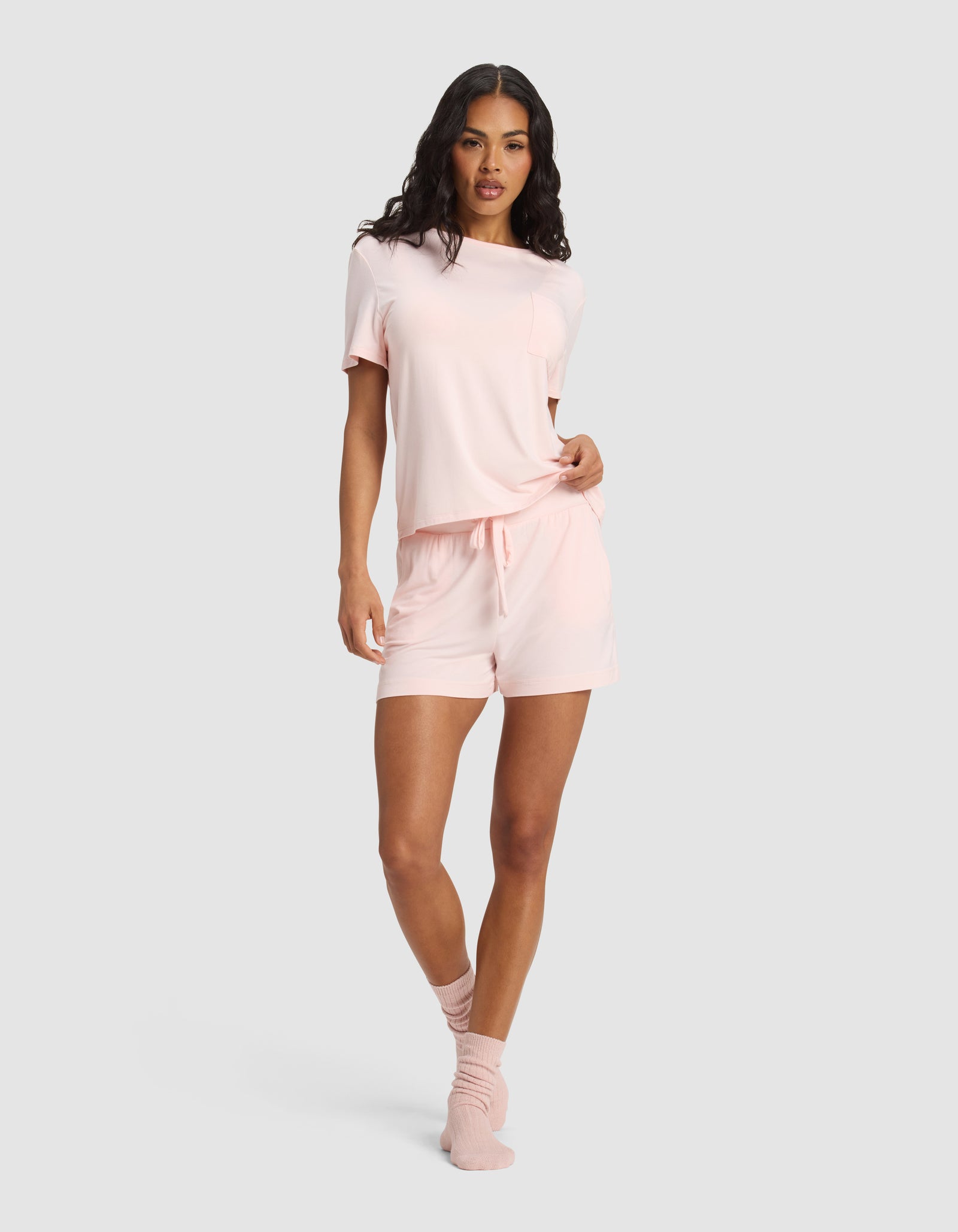 A woman stands against a plain background wearing Cozy Earth's Women's Bamboo Stretch-Knit Shorts pajama set in light pink with drawstring shorts and matching pink socks.