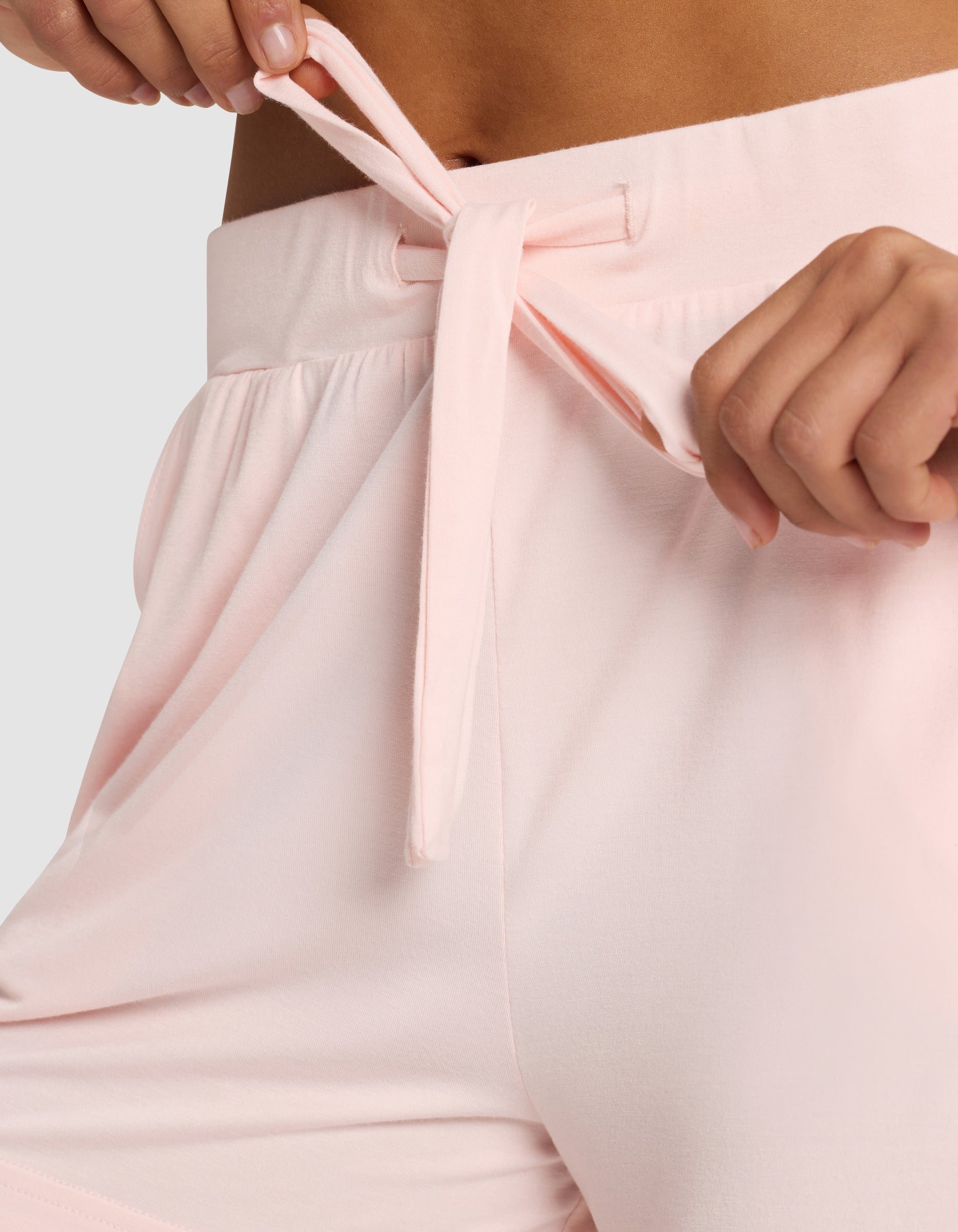 Close-up of a woman tying the drawstring on Cozy Earth's Women's Bamboo Stretch-Knit Shorts in light pink, showing her hand and part of her torso against a plain background.|Color:Blossom