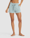 A woman, head and upper torso not visible, stands barefoot with hands in the pockets of Cozy Earth Women's Bamboo Stretch-Knit Shorts in light blue against a plain white background.|Color:Brook