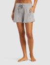 A person stands barefoot against a white background, wearing Cozy Earth's Women's Bamboo Stretch-Knit Shorts in light gray with one hand in a pocket and the other by their side.|Color:Heather Grey